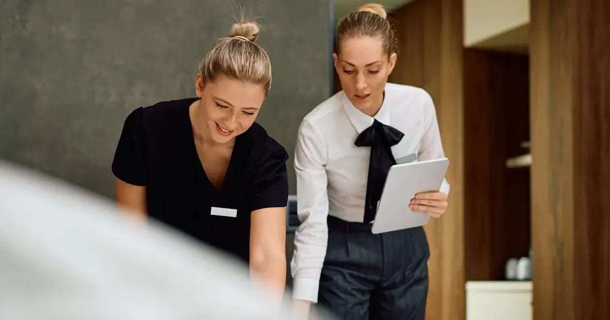 Hotel housekeepers working together in a guest room. Supervisor is holding a tablet where they can check guest departures in the RMS housekeeping app as they are entered by reception staff. 