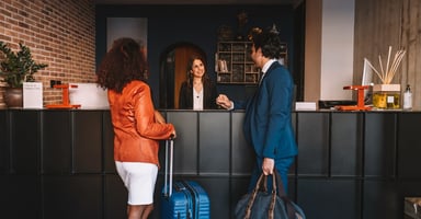 Two guests standing at a hotel reception desk with luggage while a receptionist assists them during check-in. 