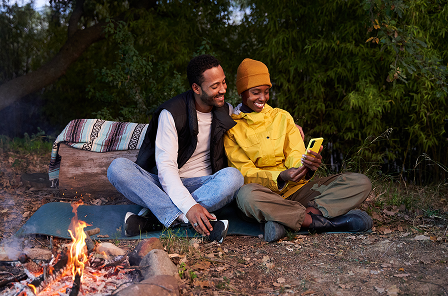 A couple outdoors, sitting next to a firecamp and looking at a phone