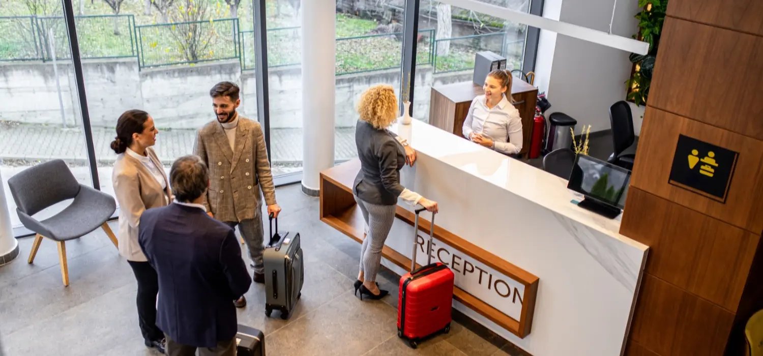 Hotel lobby with a diverse group of guests and a woman confirming her booking with the front desk staff. 