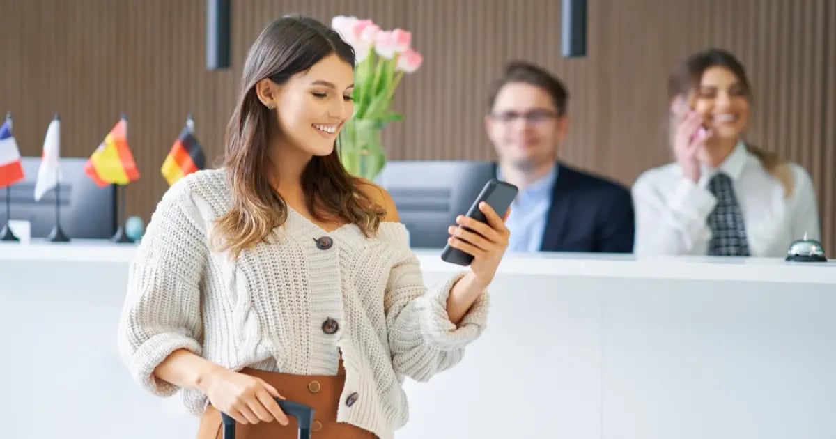 Hotel guest checking her phone at the front desk while reception staff assist in the background.