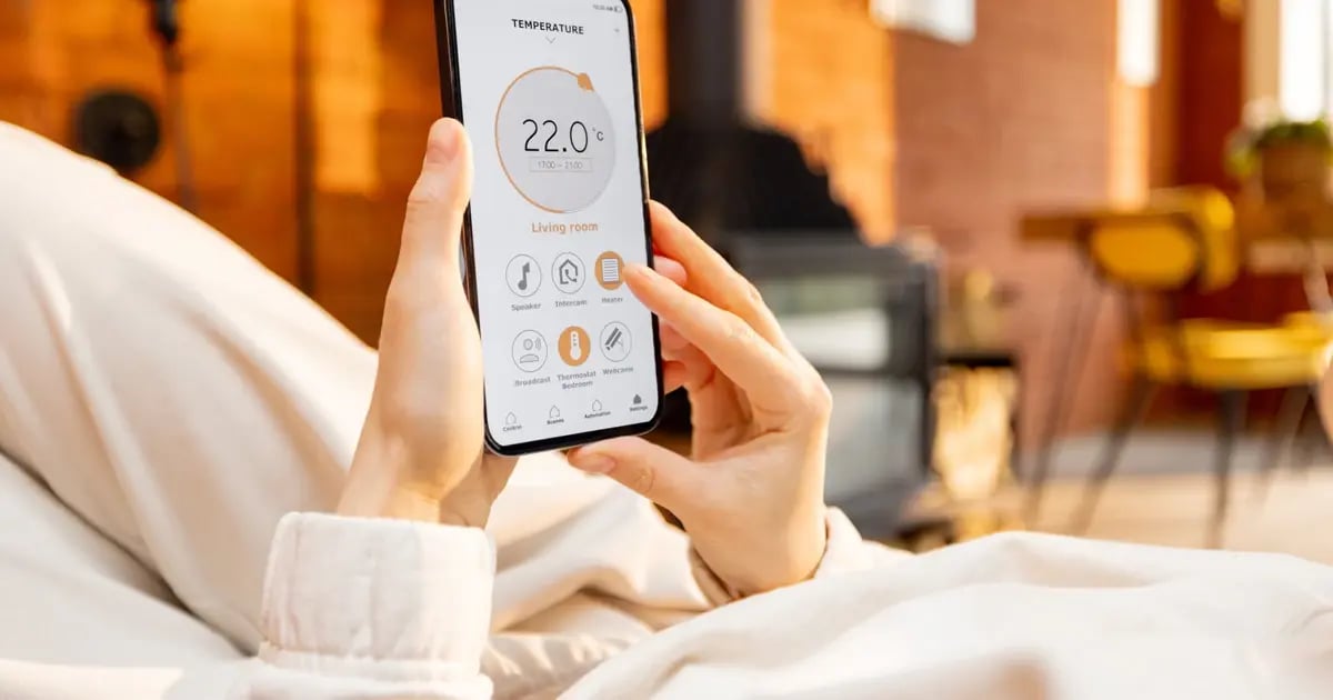 A person controlling smart home features from a smartphone while relaxing in a modern hotel-style bedroom.