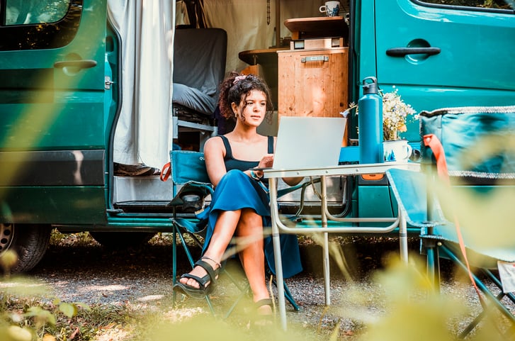 A young woman sitting at a table, working at a laptop in front of a caravan