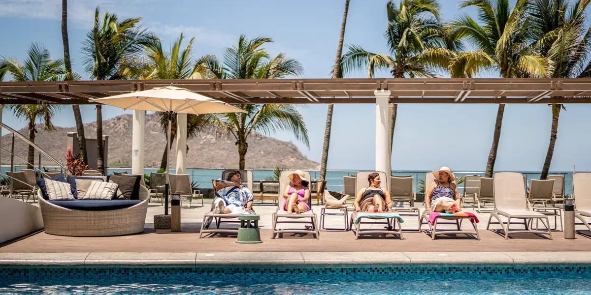 Guests lounging on sunbeds beside a resort swimming pool with palm trees and the ocean in the background on a sunny day.