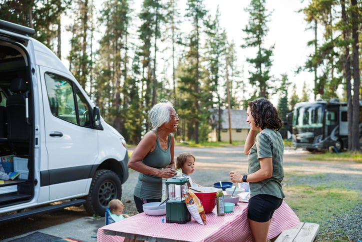 An active and healthy Pacific Islander senior woman cheerfully talks with her Eurasian adult daughter as they prepare a meal together at a picnic table at a campground while on a multi-generation family vacation.