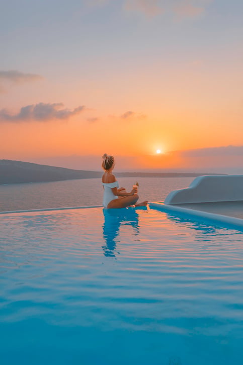 A young woman sitting by the infinity pool edge and enjoys watching sunset view and the Mediterranean sea