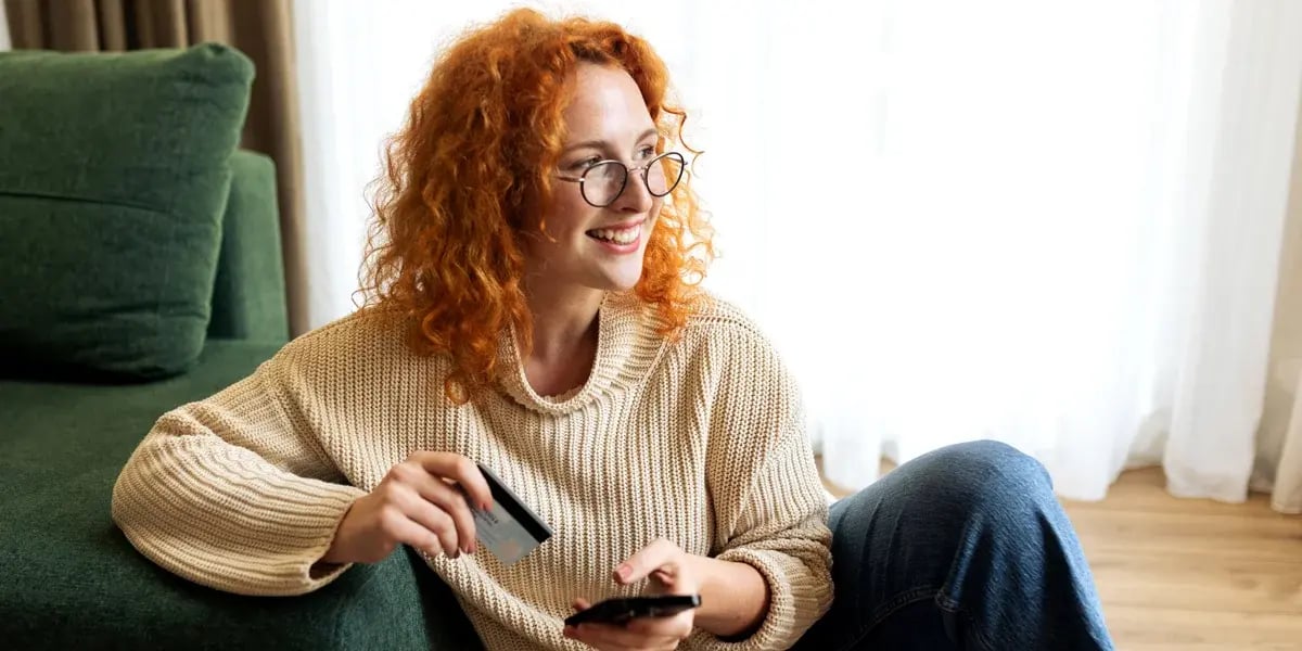 Young woman sitting on a couch at home, holding a credit card and using a laptop for online shopping. 