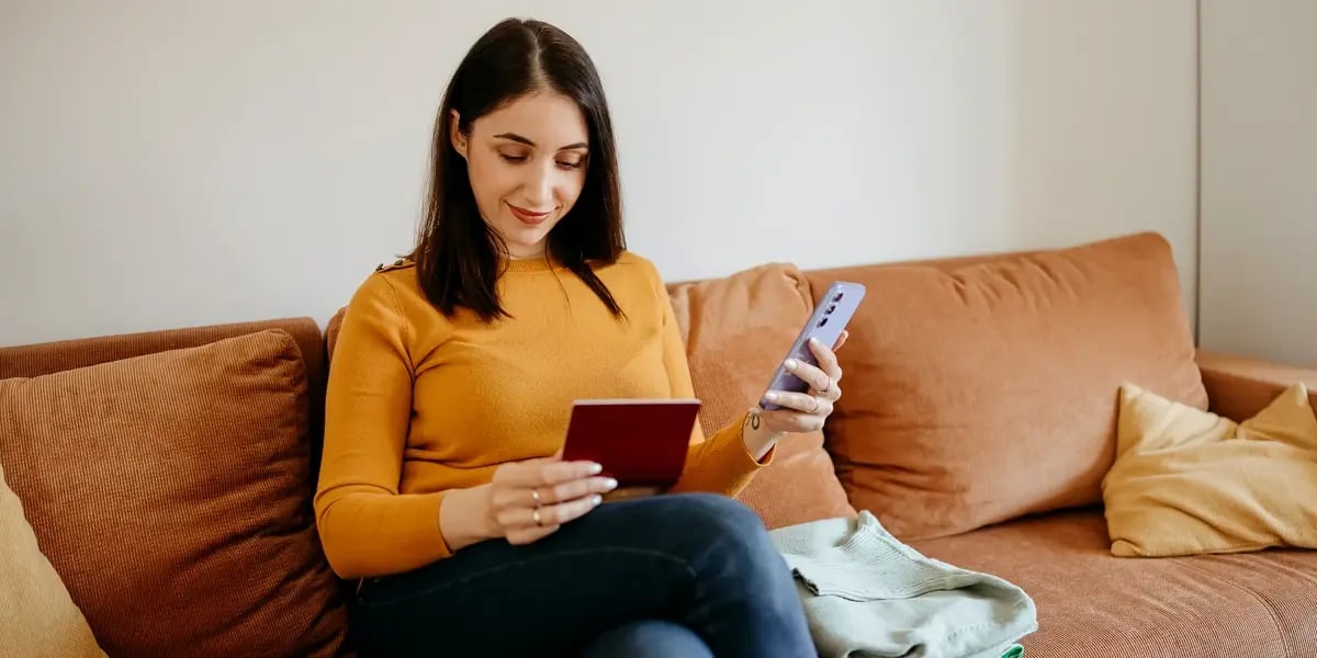 A woman in a mustard sweater sits on a brown couch holding a passport and smartphone, with folded clothes beside her and a suitcase and coffee cup nearby, suggesting she is planning or preparing for travel. 