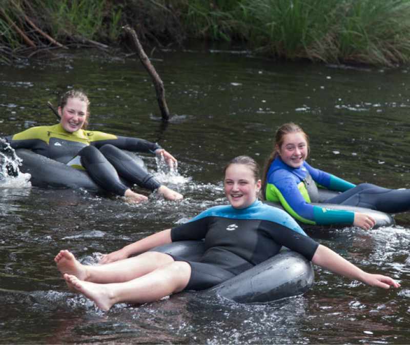Three girls in wetsuits laughing and floating on inflatable tubes in the Yarra River at Warburton Holiday Park.